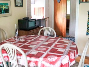 A dining area with a table covered in a red and white patterned tablecloth, white chairs, a wooden cabinet with a television, and a window with curtains. - 5 Person Holiday Home in Mellbystrand (Mellbystrand)