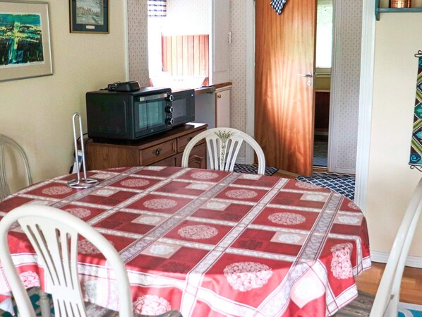 Un coin repas avec une table recouverte d’une nappe à motifs rouges et blancs, des chaises blanches, un meuble en bois avec un téléviseur et une fenêtre avec des rideaux.