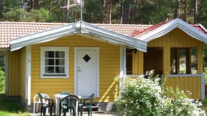 A yellow cabin with a red-tiled roof, a white door, and a small porch with outdoor furniture.