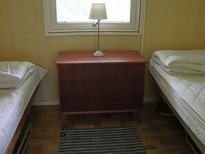 Um quarto pequeno com uma mesa de cabeceira de madeira, uma luminária, duas camas com roupa de cama branca e um tapete listrado no chão.