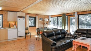 A cozy cabin interior with a wooden floor, a black leather sofa, a dining table, and a kitchen area with a refrigerator and cabinets.