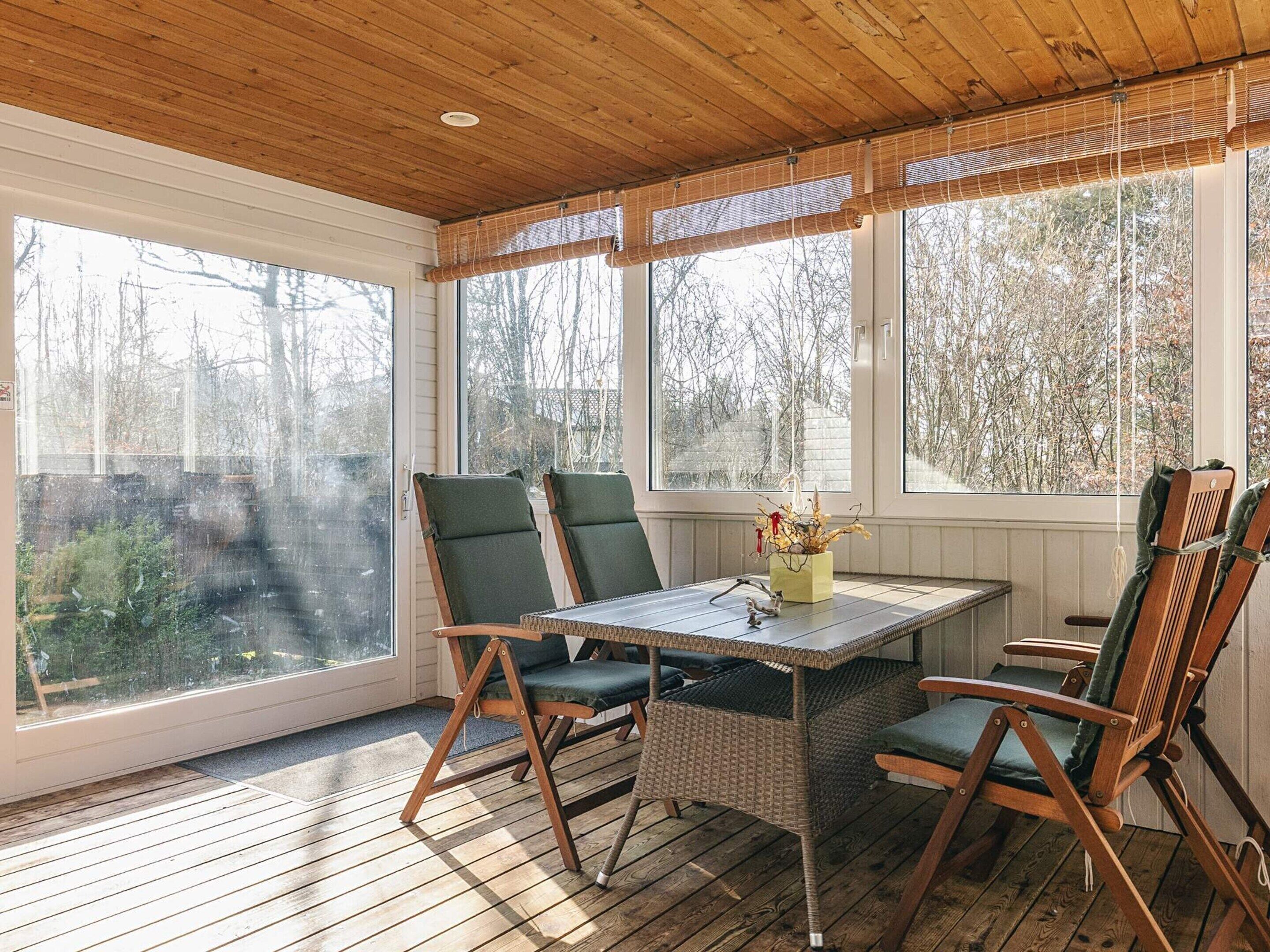 Een overdekt terras met een tafel en stoelen, grote ramen en een houten plafond.