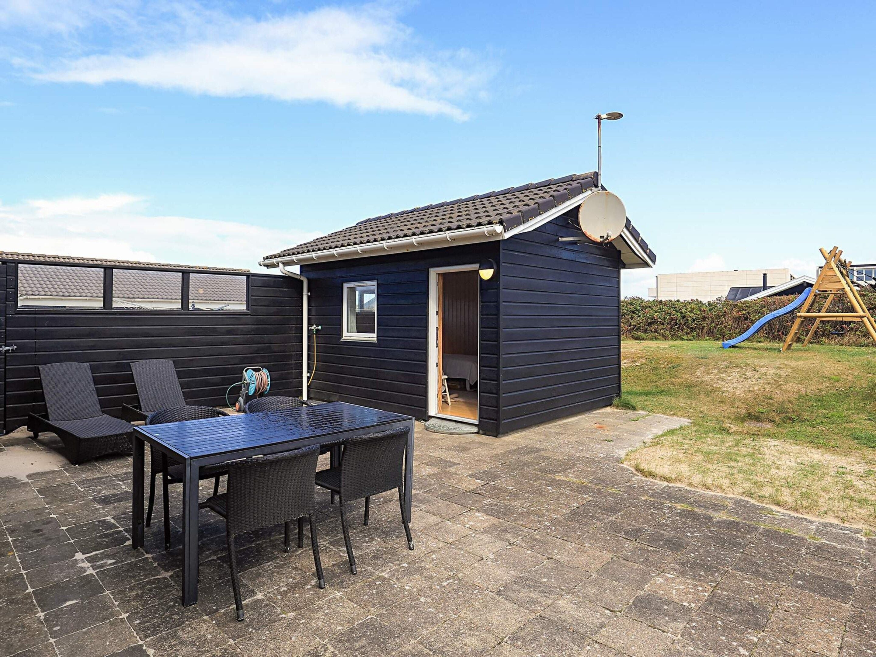 En terrasse med et bord og stole, et lille skur med dør, og en rutchebane i baggrunden.
