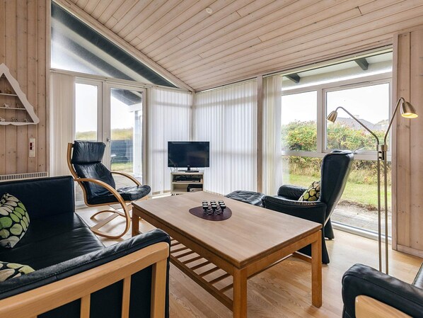 A cozy living room with a wooden table, leather chairs, a television, and large windows offering a view of the outdoors.