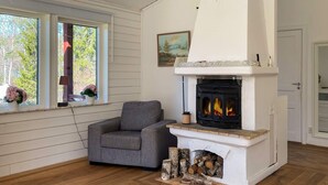 Une pièce avec une cheminée, un fauteuil gris, un plancher en bois et une fenêtre donnant sur les arbres.