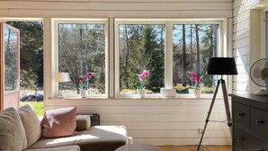 Ein gemütliches Zimmer mit einer Couch, einem kleinen runden Tisch und einer Kommode. Drei Fenster mit Blumentöpfen auf der Fensterbank.