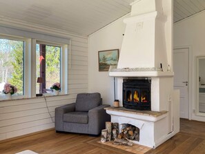 A room with a fireplace, a gray armchair, wooden flooring, and a window with a view of trees.
