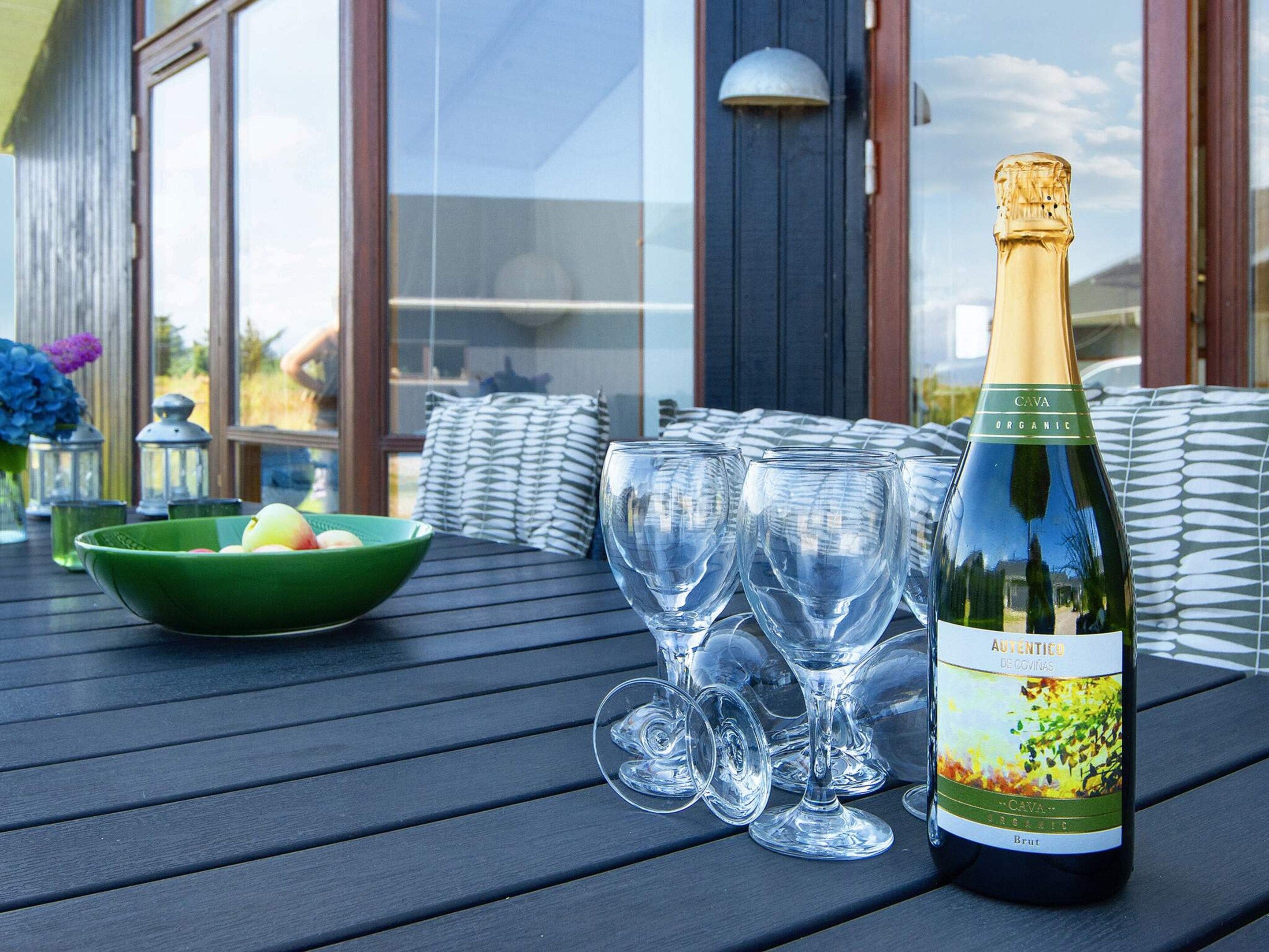 A bottle of Cava Organic wine, several wine glasses, a bowl of fruit, and a green bowl on a wooden table.