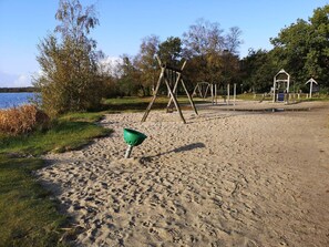 A sandy beach with a green bucket, swings, and a small structure near a body of water.