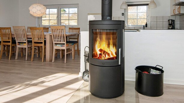 A modern living room with a wood-burning stove, a dining area with wooden chairs, and a kitchen with a coffee maker. - Tranquil Holiday Home in Ulfborg With Sauna (Ulfborg)