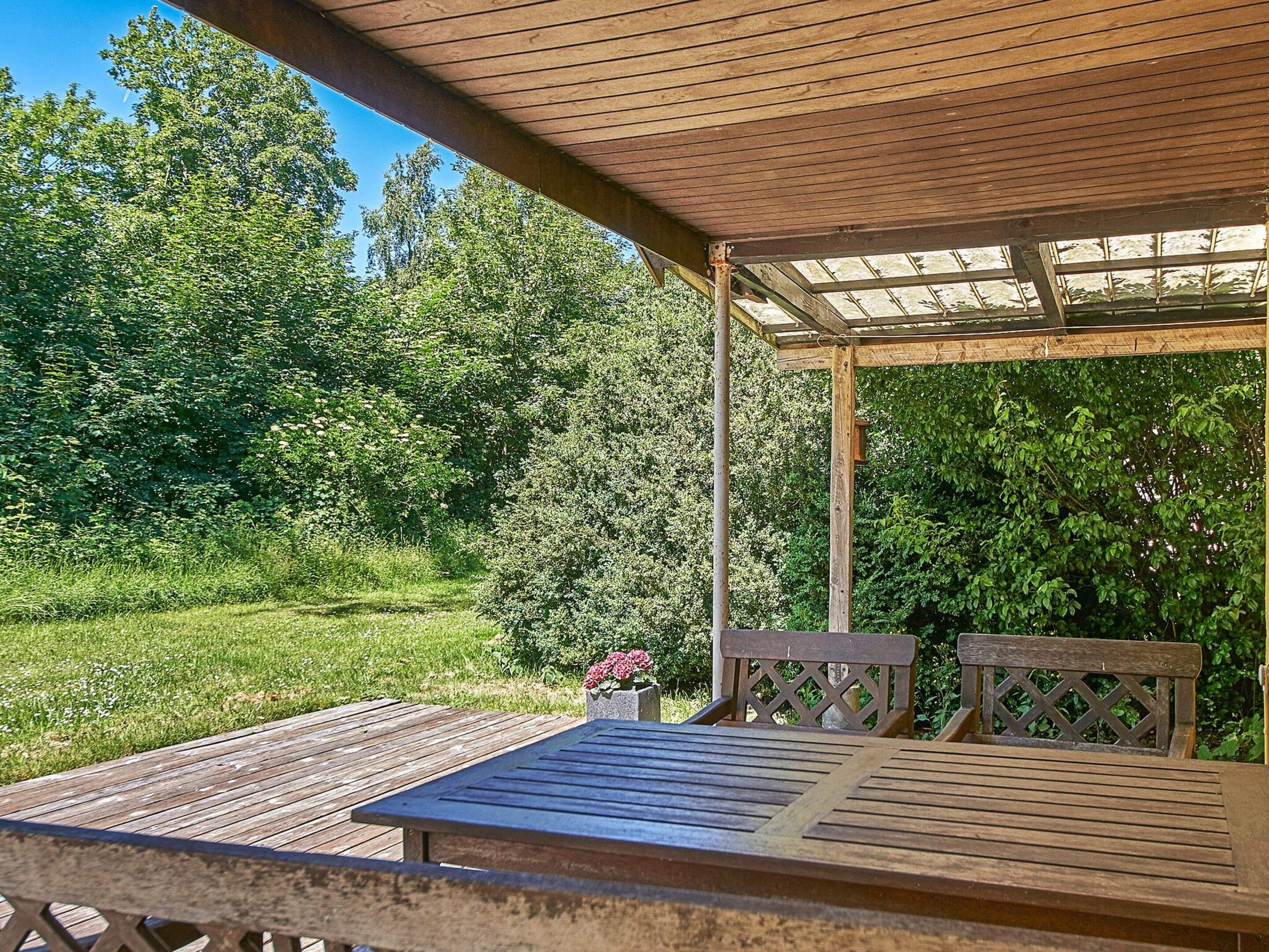 An outdoor wooden deck with a pergola, surrounded by lush greenery and trees.