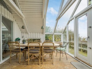 A covered patio with a table and chairs, large windows, and a view of greenery.