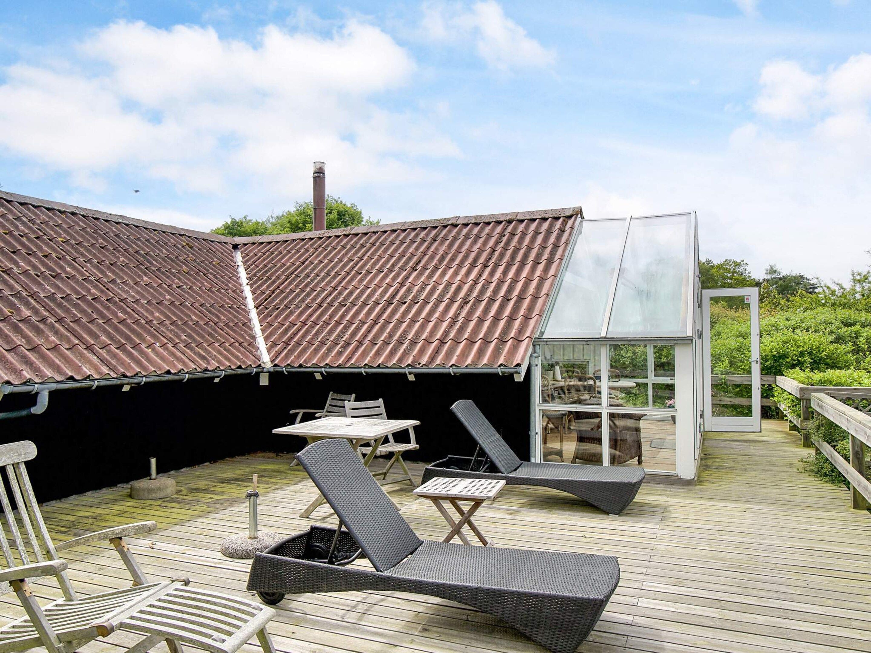 A covered outdoor area with a wooden deck, lounge chairs, and a glass-enclosed structure.