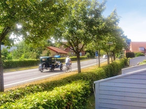 Une rue bordée d’arbres et de bâtiments résidentiels, où l’on aperçoit une voiture ancienne et un cycliste.