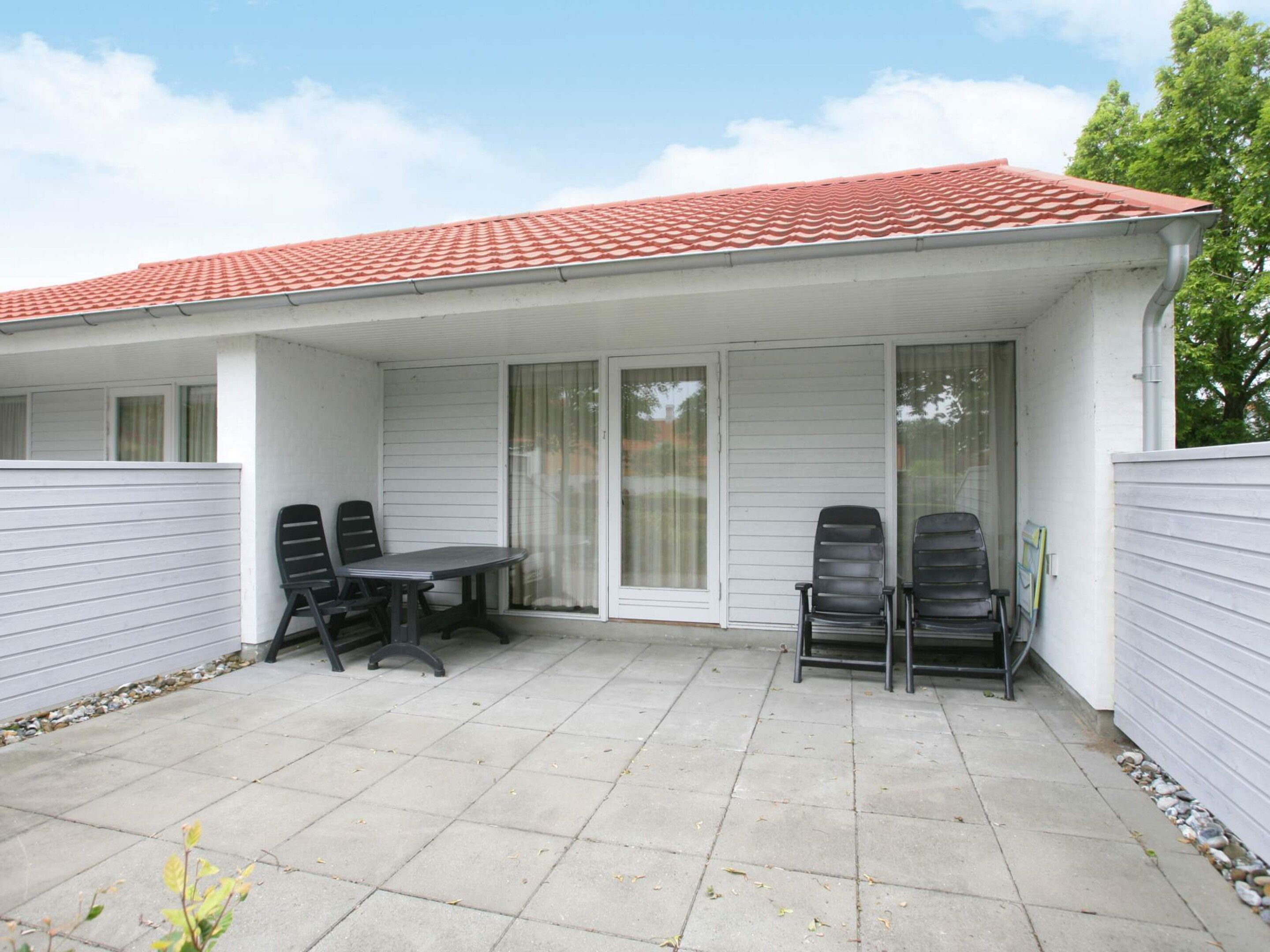A covered patio area with a table and four chairs, a sliding glass door, and a red-tiled roof.