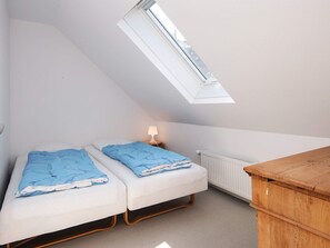 Una habitación pequeña con techo inclinado, dos camas, un aparador de madera, un radiador y un tragaluz.