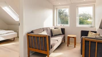 A modern, well-lit room with a sofa, a chair, and a small table. There are two windows providing natural light and a view of greenery outside.