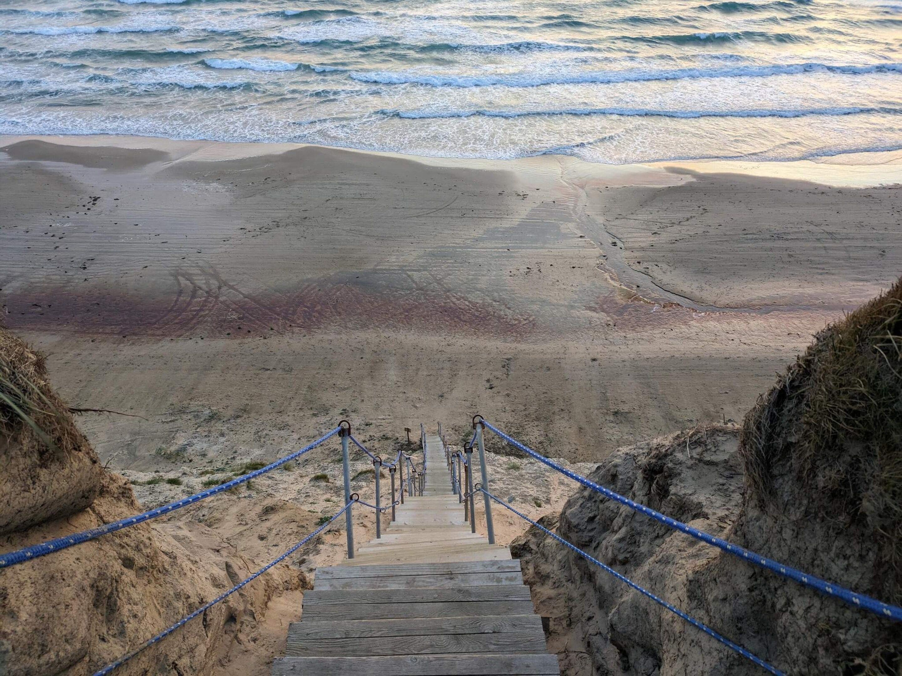 A wooden staircase leading down to a beach with a sandy shore and ocean waves in the background.