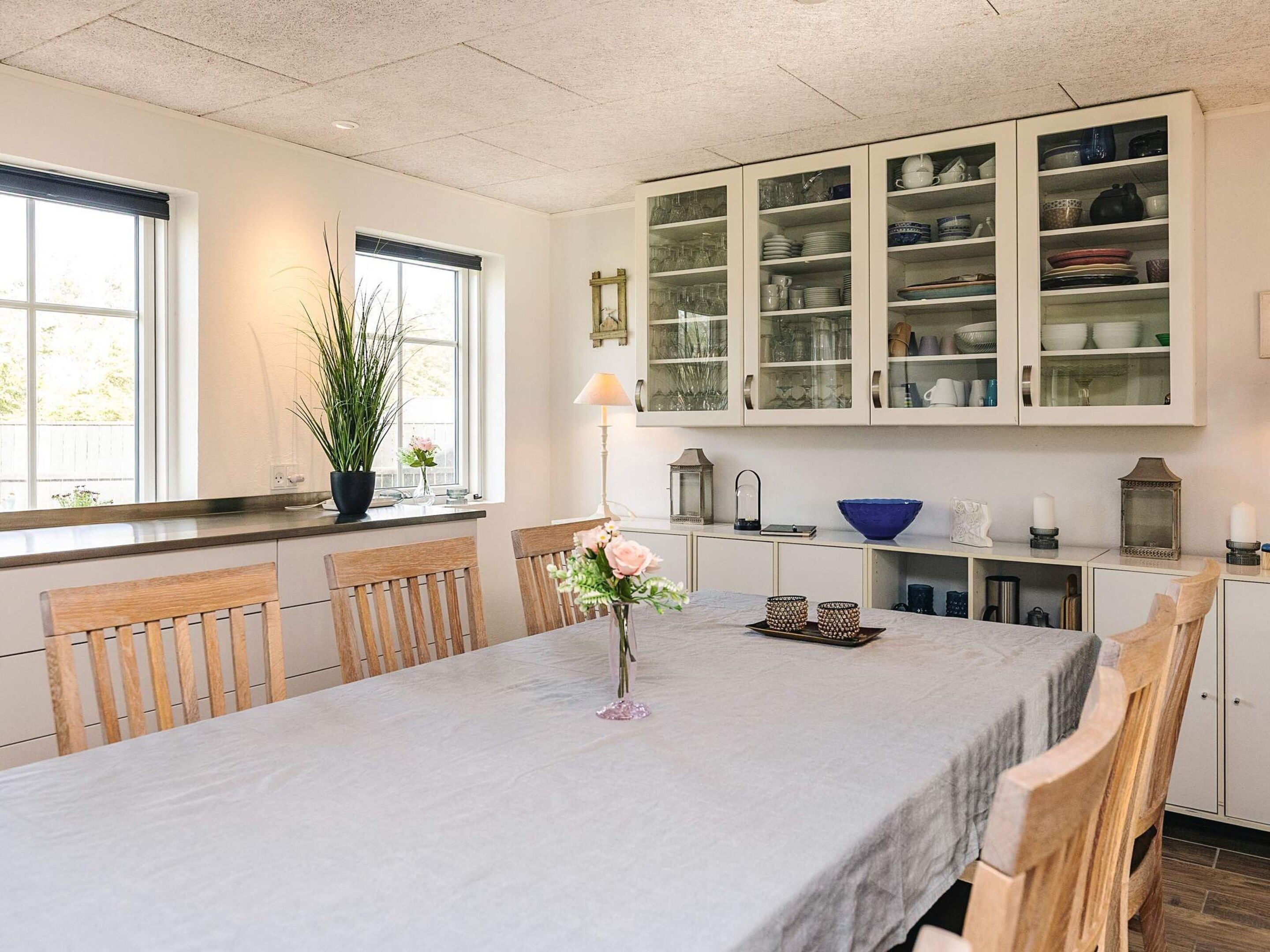 A dining area with a table set for four, surrounded by wooden chairs. A vase with flowers is placed on the table.