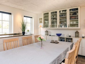 Uma sala de jantar com uma mesa posta para quatro pessoas, rodeada por cadeiras de madeira. Um vaso com flores está sobre a mesa.