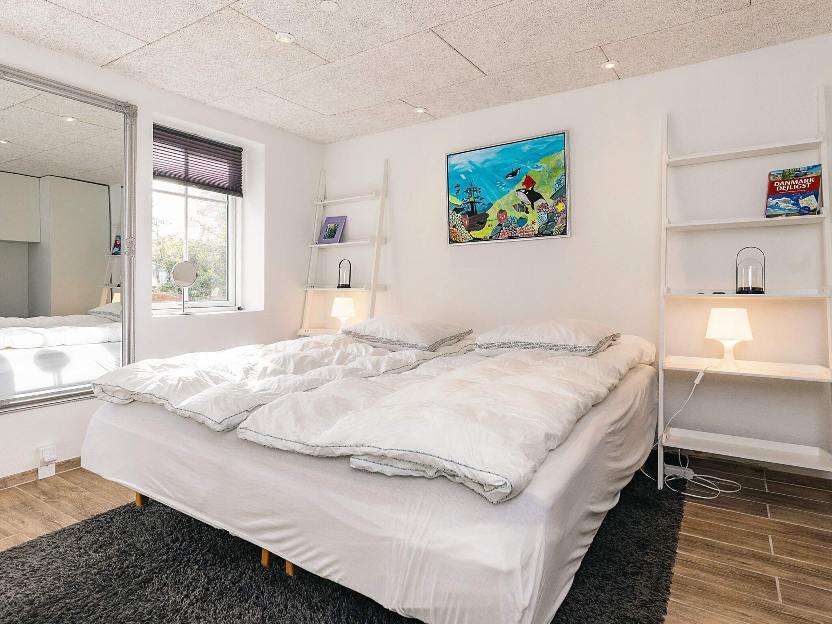 A modern bedroom with a large bed, bedside lamps, a window with blinds, a painting on the wall, and a shelf with a book and a lamp.