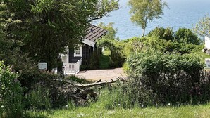 A small cabin with a wooden deck, surrounded by dense greenery and overlooking a body of water. - 6 Person Holiday Home in Hasle (Hasle)