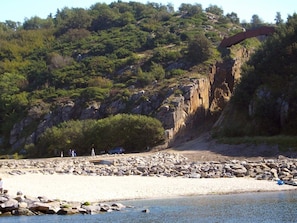 Uma praia com terreno rochoso, céu claro e uma ponte ao fundo.