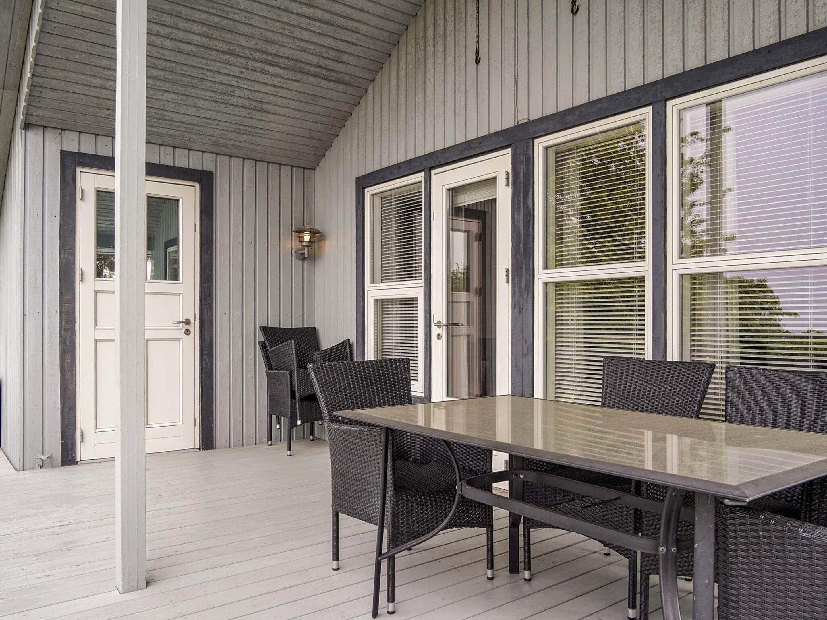 Een overdekt terras met een tafel, stoelen, een deur en ramen met zonwering.