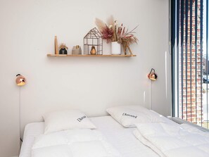 A bedroom with a bed, two bedside lamps, a shelf with decorative items, and a window with a view of a brick wall.