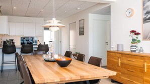 Un comedor moderno con una mesa de madera, sillas negras, una lámpara colgante en el centro y una cocina al fondo.