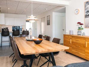 A modern dining area with a wooden table, black chairs, a central light fixture, and a kitchen in the background. - 5 Star Holiday Home in Ringkobing (Ringkobing)