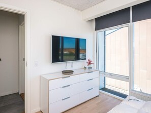 A modern bedroom with a large window, a white dresser with drawers, a flat-screen TV mounted on the wall, and a small plant on the dresser.
