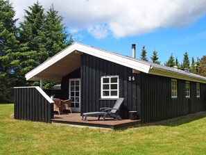 A black wooden cabin with a porch, number 54, featuring a single window, a chimney, and a small deck with outdoor furniture. - 4 Star Holiday Home in Blokhus (Blokhus)