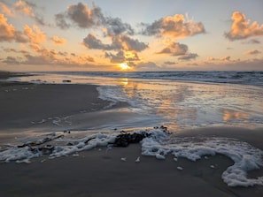 A beach scene with the sun setting over the ocean, waves gently lapping at the shore, and scattered rocks along the sand.