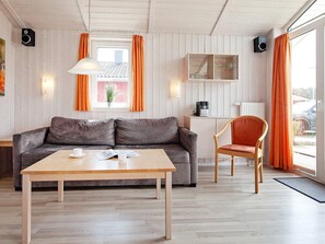 Ein modernes Wohnzimmer mit einer Couch, einem Holztisch und einem Sessel. Ein Fenster mit Vorhängen, ein Wandlautsprecher und eine Heizung sind ebenfalls zu sehen.