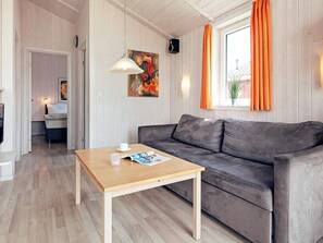 A room with a wooden floor, a grey sofa, a wooden coffee table, a painting on the wall, and a window with orange curtains.