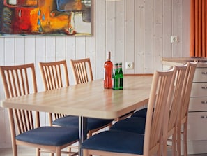 A dining area with a wooden table and chairs, a painting on the wall, and a kitchen area with cabinets and a countertop. - Holiday Home in GrÃ¶mmitz (Groemitz)