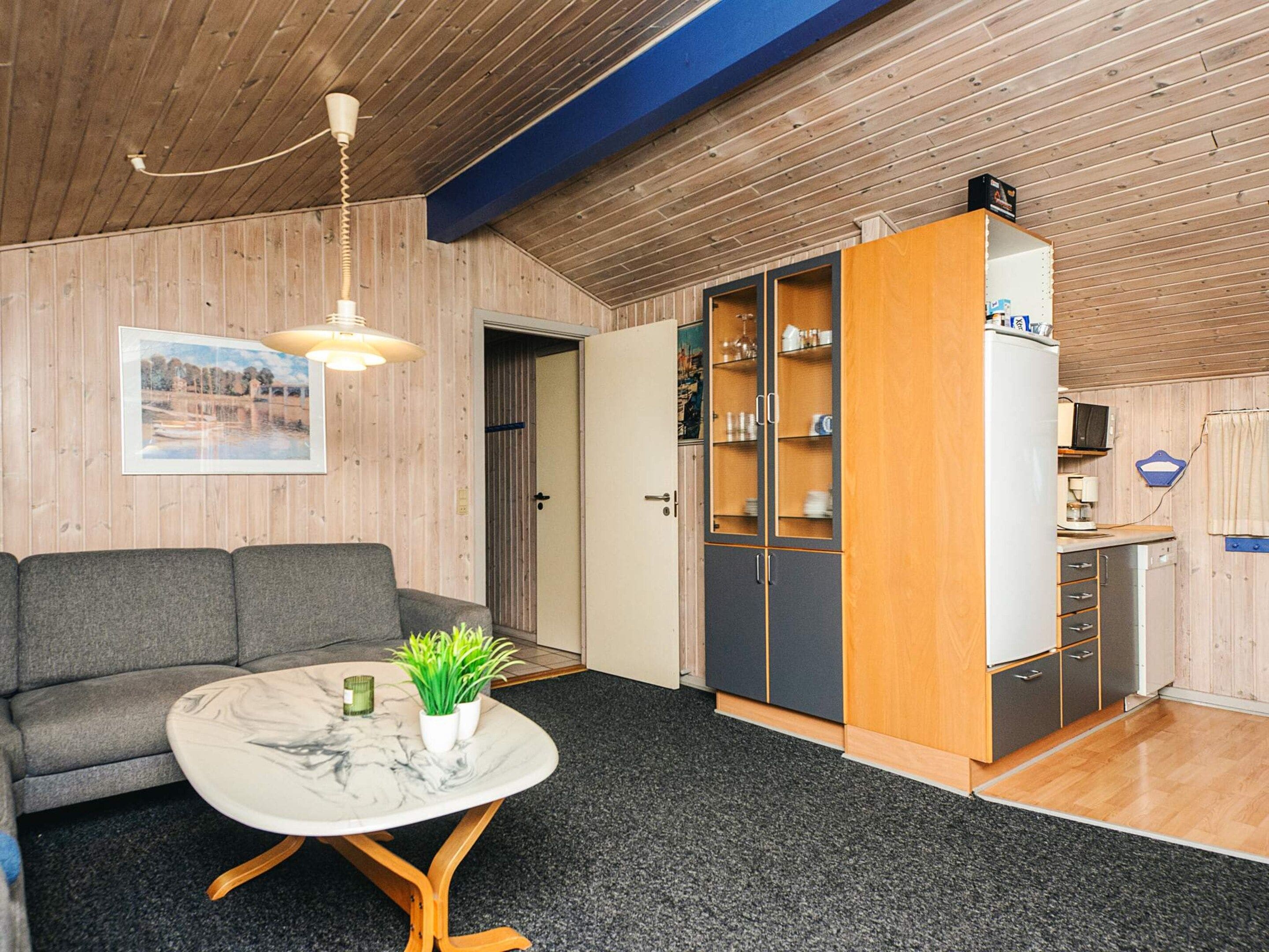 Un espacio habitable compacto con sofá, una mesa de comedor con planta, zona de cocina con armarios y techo con paneles de madera.