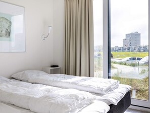 A hotel room with a bed, a bedside table, a lamp, and a window with a view of buildings and a parking lot. - Holiday Home in Wendtorf (Schönberg)