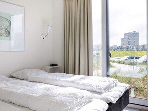 A hotel room with a bed, a bedside table, a wall-mounted lamp, and a window with a view of a parking lot and buildings.