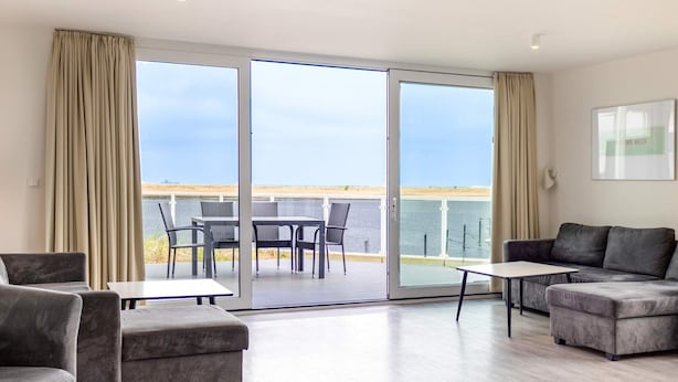 A modern living room with a grey sofa, a small table, and a balcony with a view of the sea. - Holiday Home in Wendtorf (Schönberg)