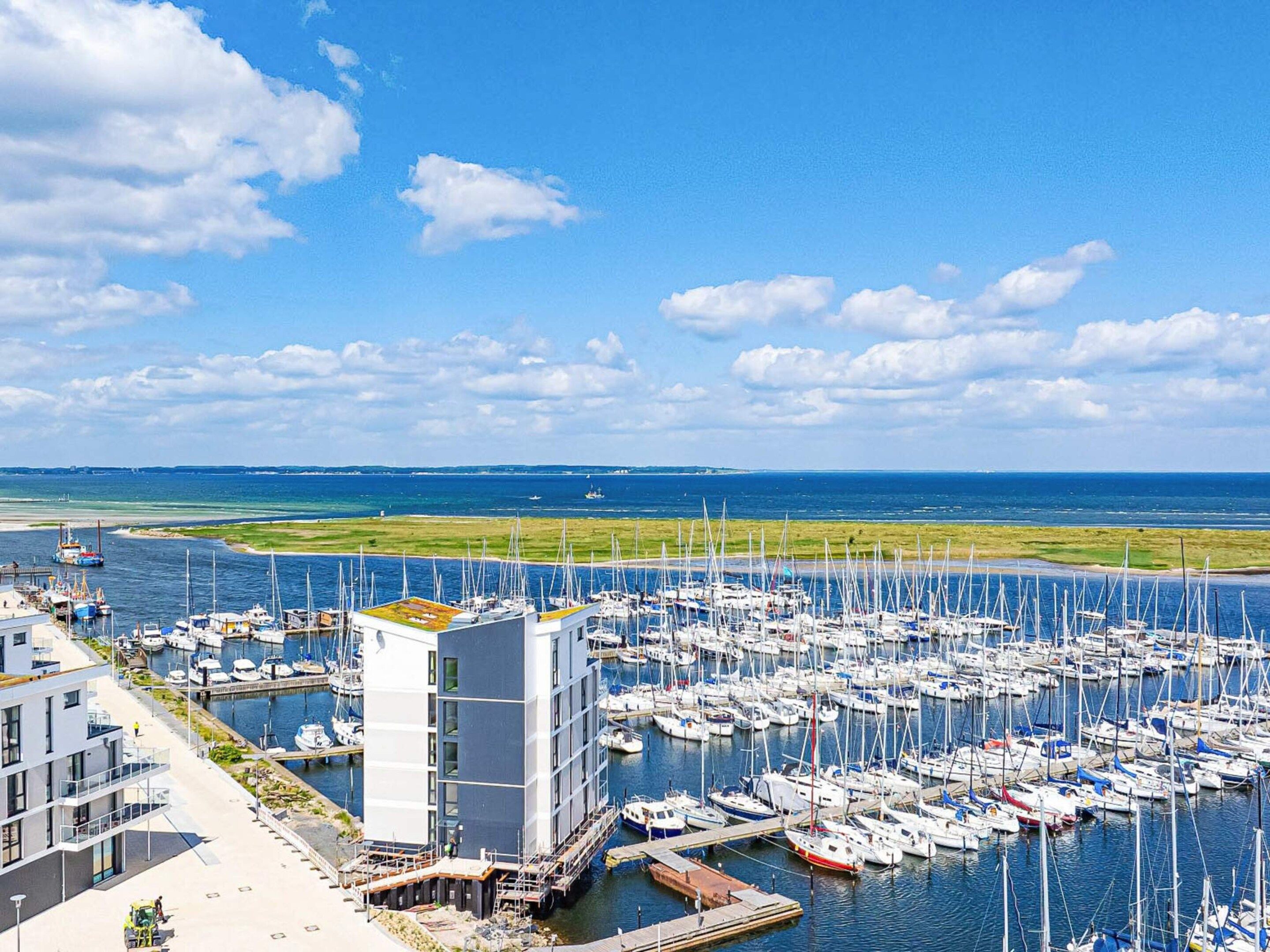 Un porto turistico con numerose imbarcazioni ormeggiate, un edificio moderno a tetto piatto e una vista aperta su mare e cielo.