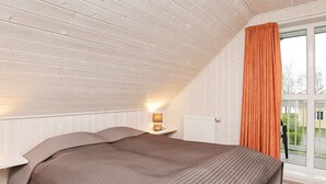 A bedroom with a sloped ceiling, a bed with a brown quilt, a bedside lamp, and a window with an orange curtain.