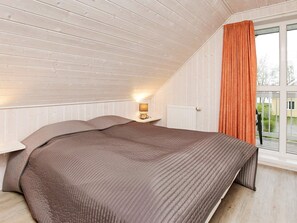 A bedroom with a sloped ceiling, a bed with a brown quilt, a bedside lamp, and a window with an orange curtain.