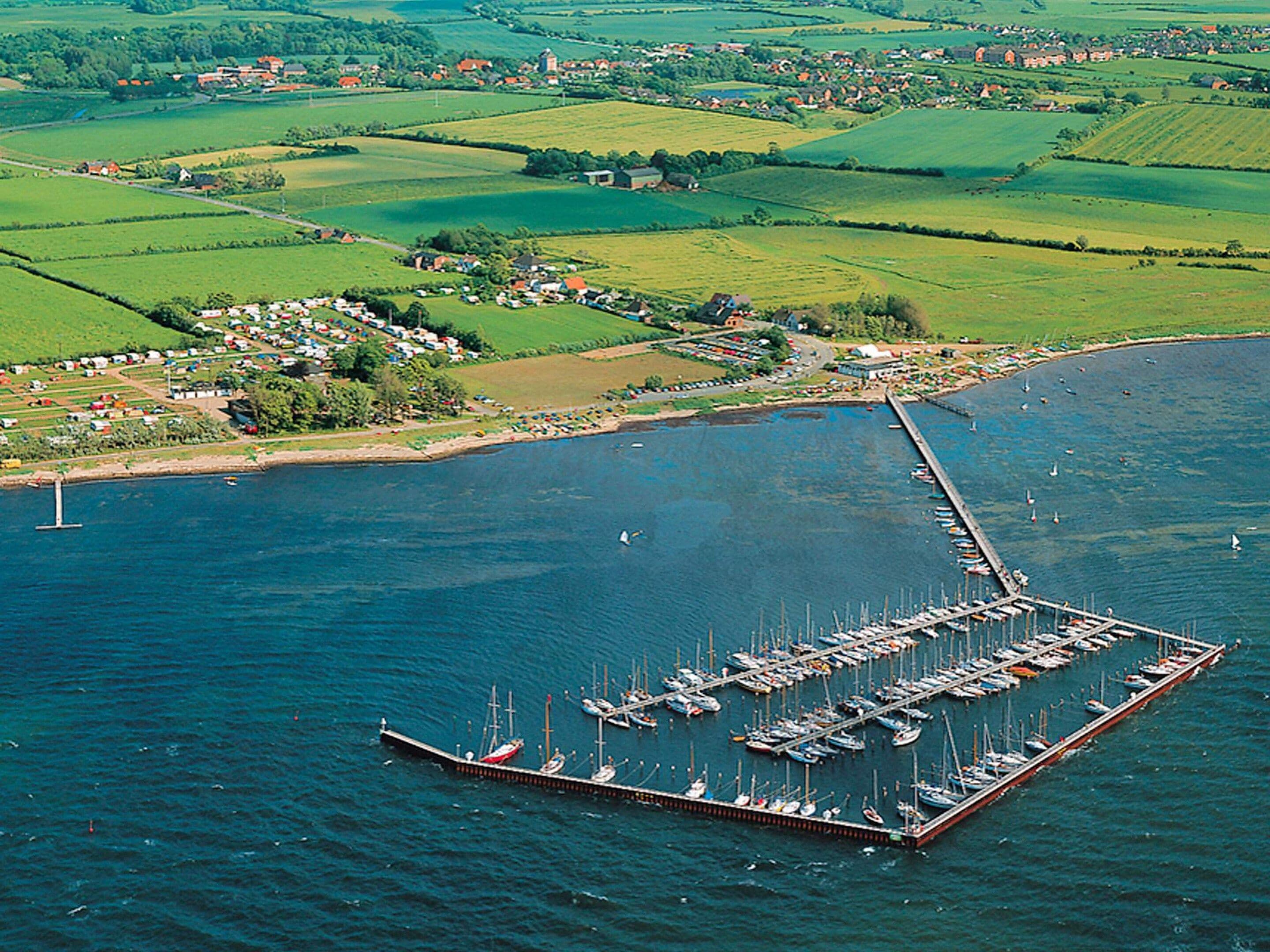 An aerial view of a marina with numerous boats docked, surrounded by green fields and a small village.