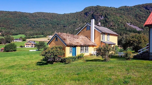 Ein zweistöckiges Haus mit einem Strohdach, umgeben von einer grünen Wiese und einem bewaldeten Hügel.