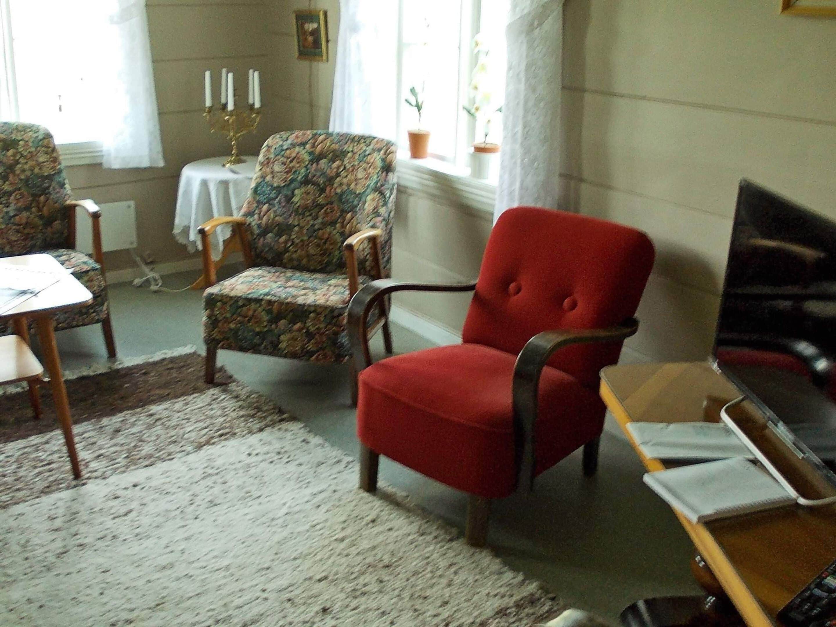 Ein Zimmer mit einem roten Sessel, einem floralen Sessel, einem kleinen Tisch mit einem Kandelaber und einem Blumentopf am Fenster.