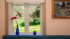 Ein Zimmer mit großem Fenster, einem Gemälde an der Wand und einer blauen Vase mit Blumen auf dem Fensterbrett.