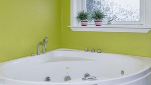 Uma banheira branca com etiqueta Camargue, uma janela com padrão floral e duas plantas em vasos sobre uma estante.