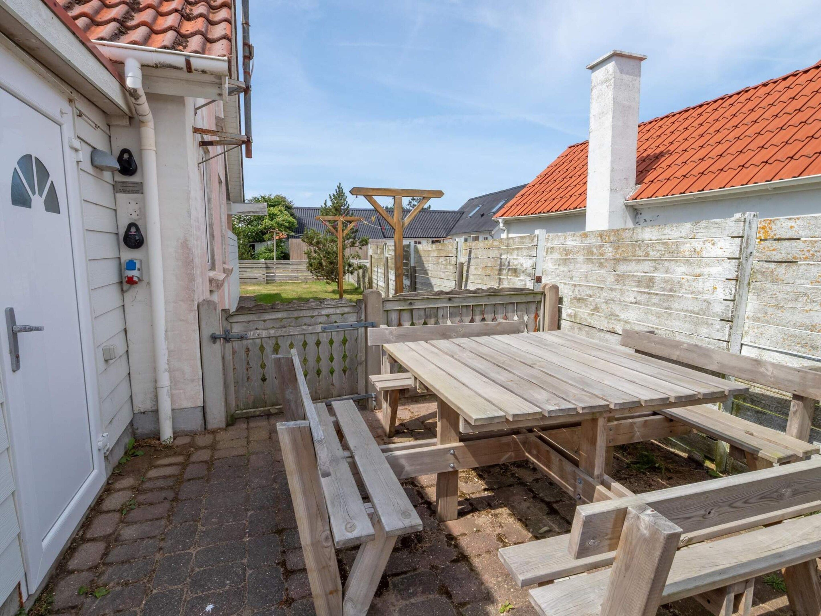 An outdoor patio area with a wooden table and benches, a white door with a security device, and a red-tiled roof visible in the background.